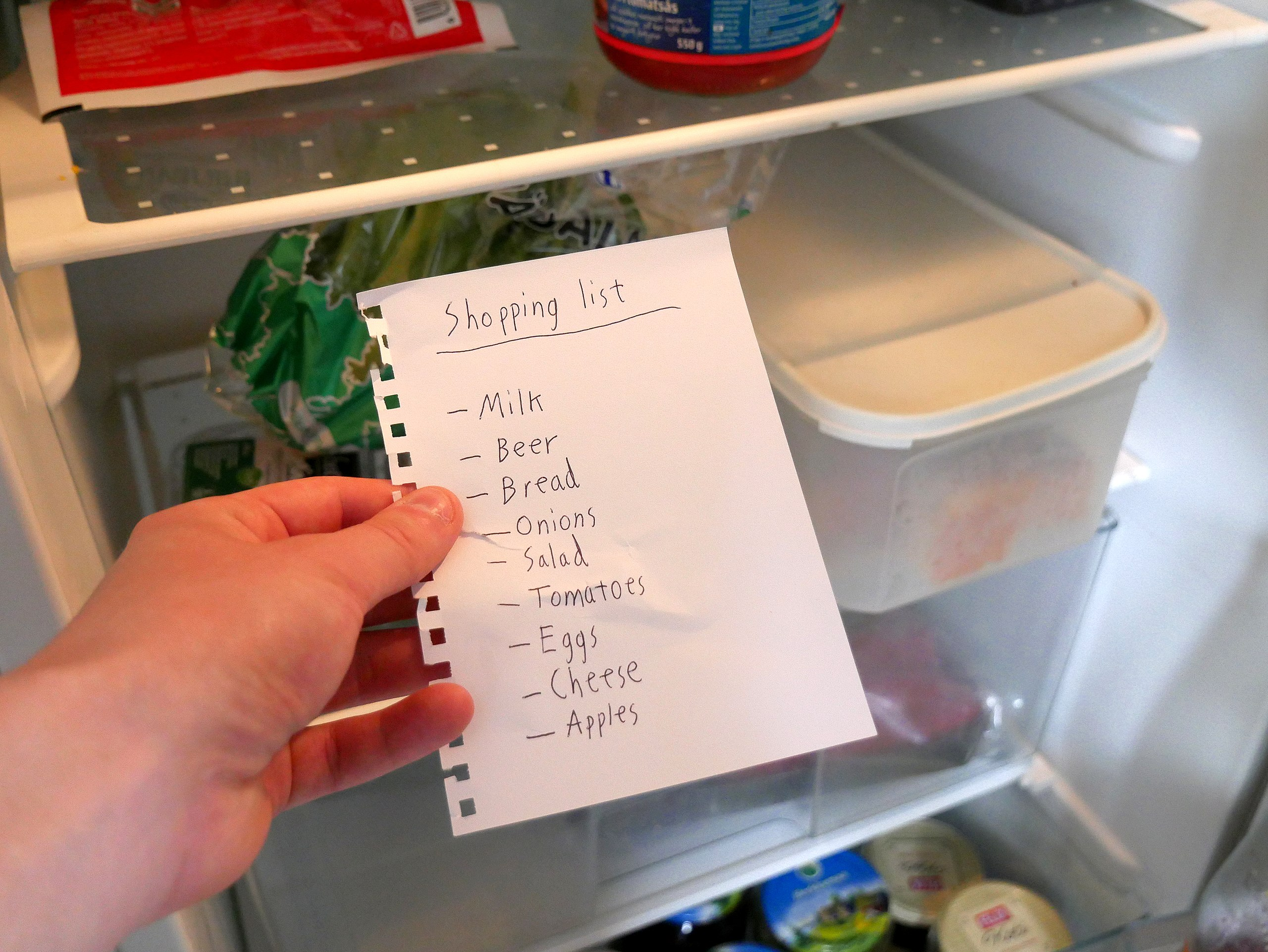 Left Hand Holding a Short Grocery List With Opened Refrigerator in the Background