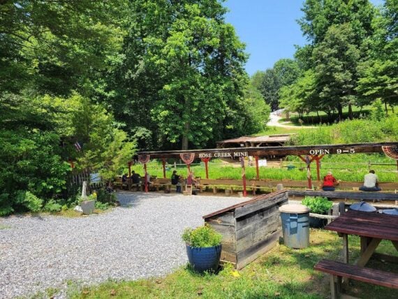 People sit at long benches under a roof marked “Rose Creek Mine” and “Open 9-5” in a wooded area.
