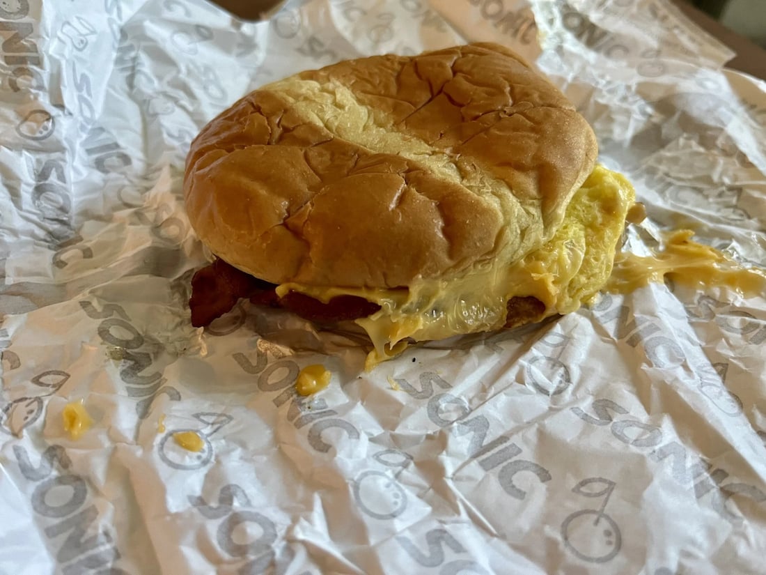 A breakfast sandwich with a slightly squished bun, melted cheese, egg, and bacon sits on Sonic-branded wrapper paper—a classic fast-food breakfast enjoyed during early fast-food breakfast hours.
