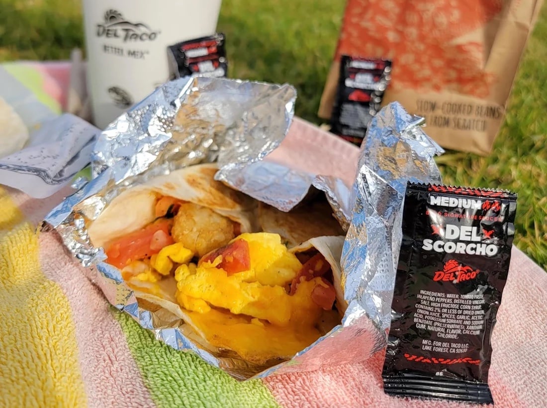 A breakfast burrito with eggs, sausage, and tomatoes is unwrapped on a picnic blanket, surrounded by Del Taco hot sauce packets—perfect for enjoying fast-food breakfast hours outdoors with a drink cup and brown paper bag.