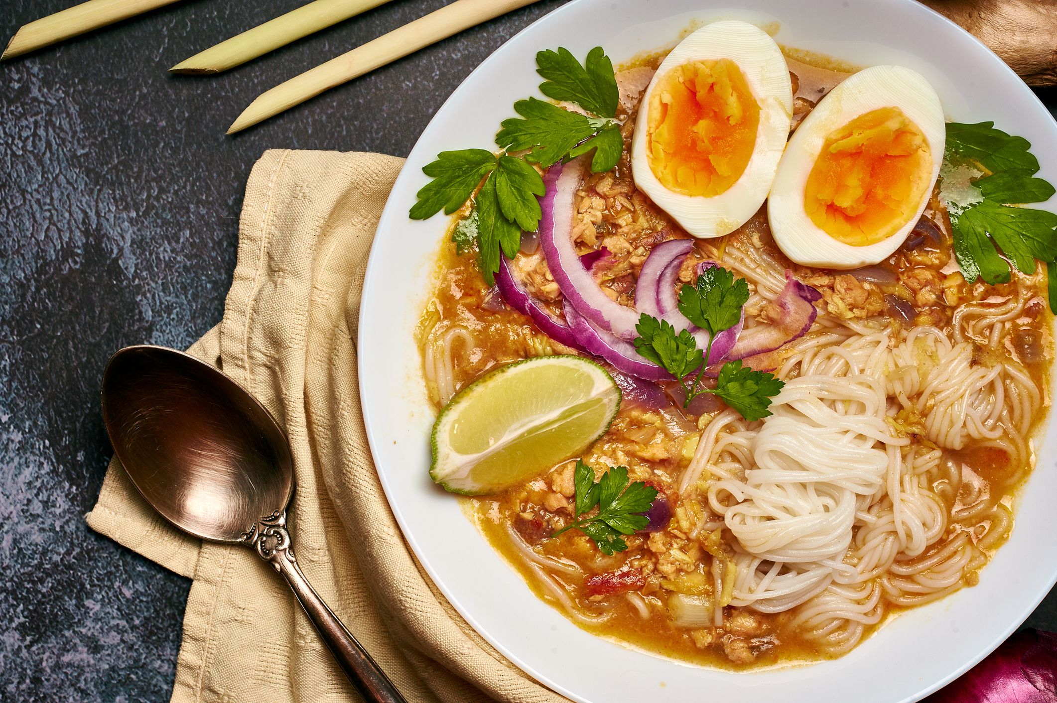 Mohinga at dark background in white plate. Mohinga is a burmese cuisine fish soup with catfish, rice noodles, chicken eggs and lime.