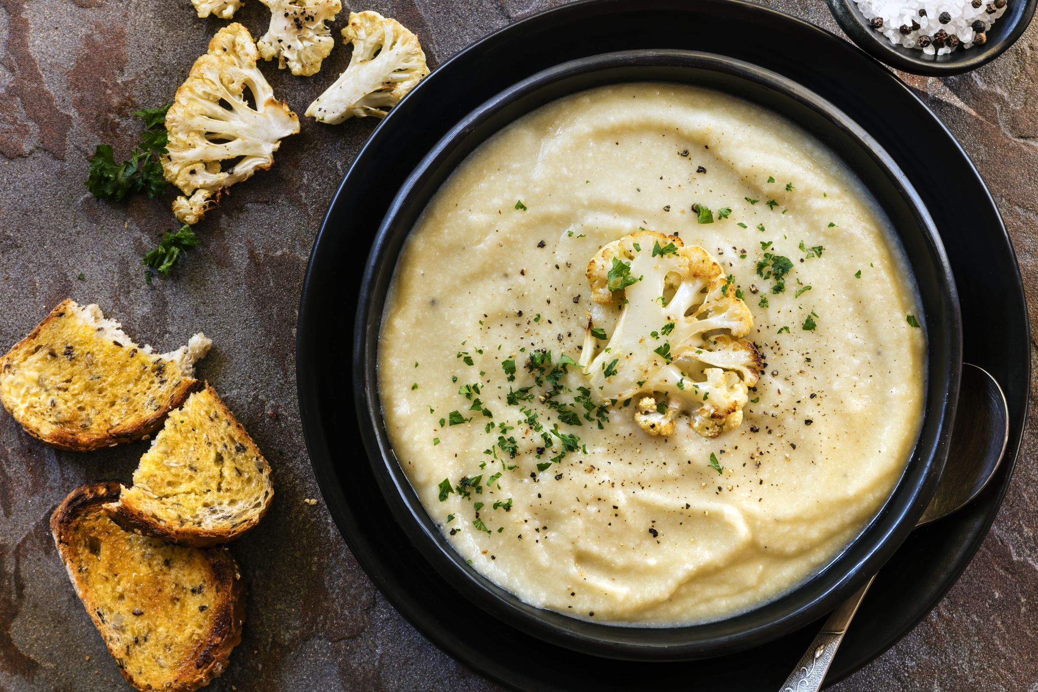 Roasted cauliflower soup in rustic black bowl, with crusty sourdough toast. Top view.