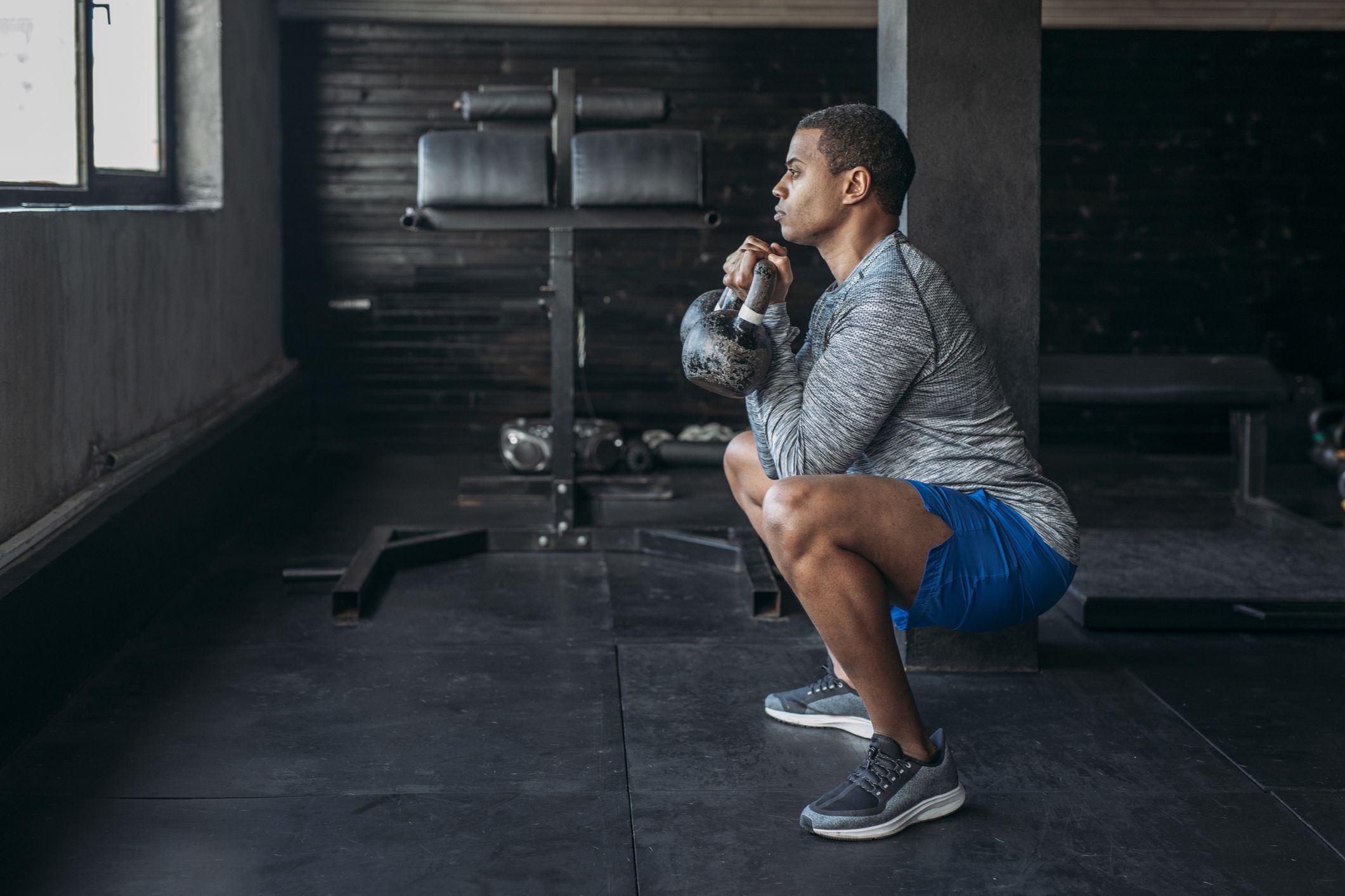 Handsome serious African sportsman doing a squat with two kettlebells in his arms.