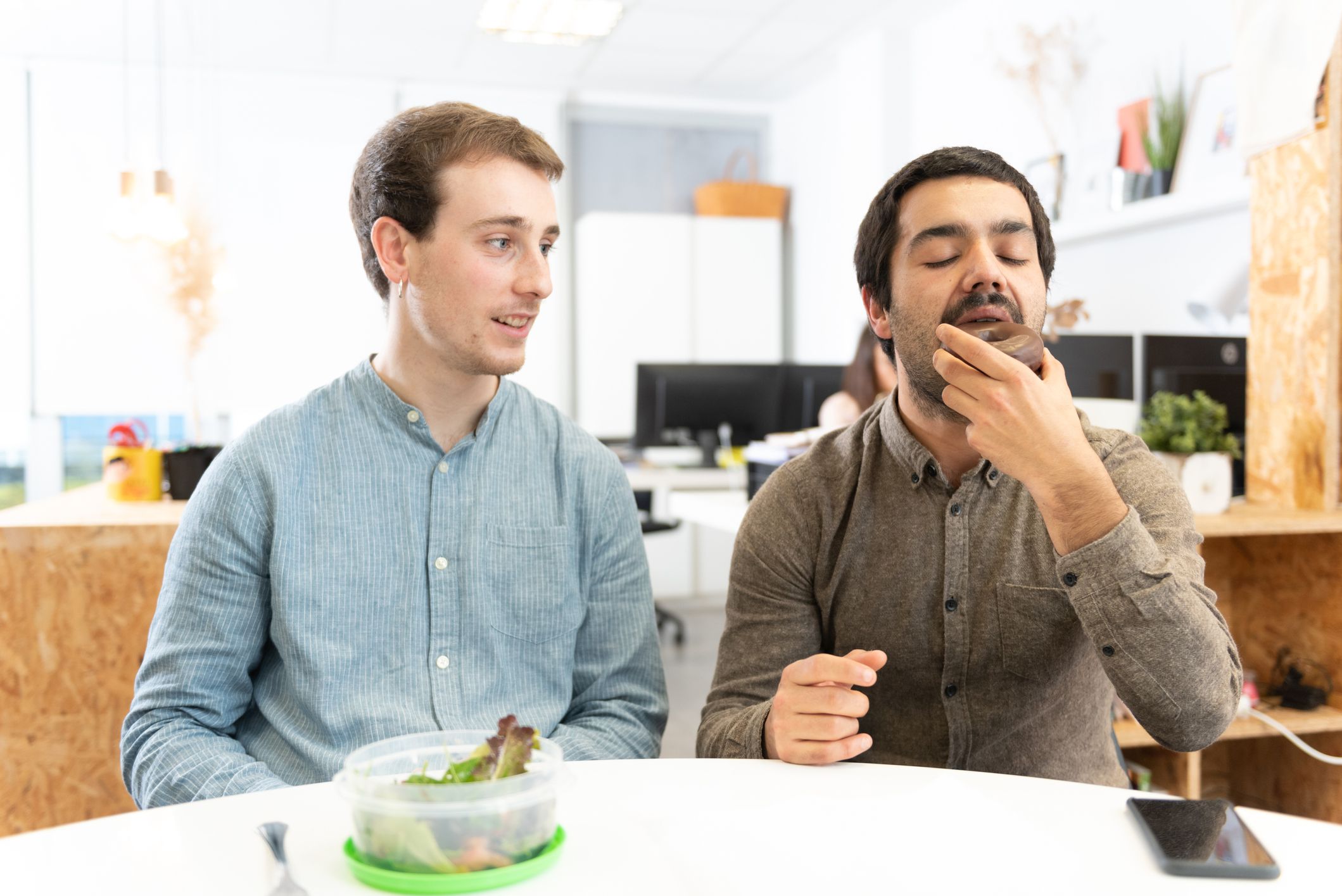 A man with a salad looking with envy to his coworker eating a sweet food in the office.