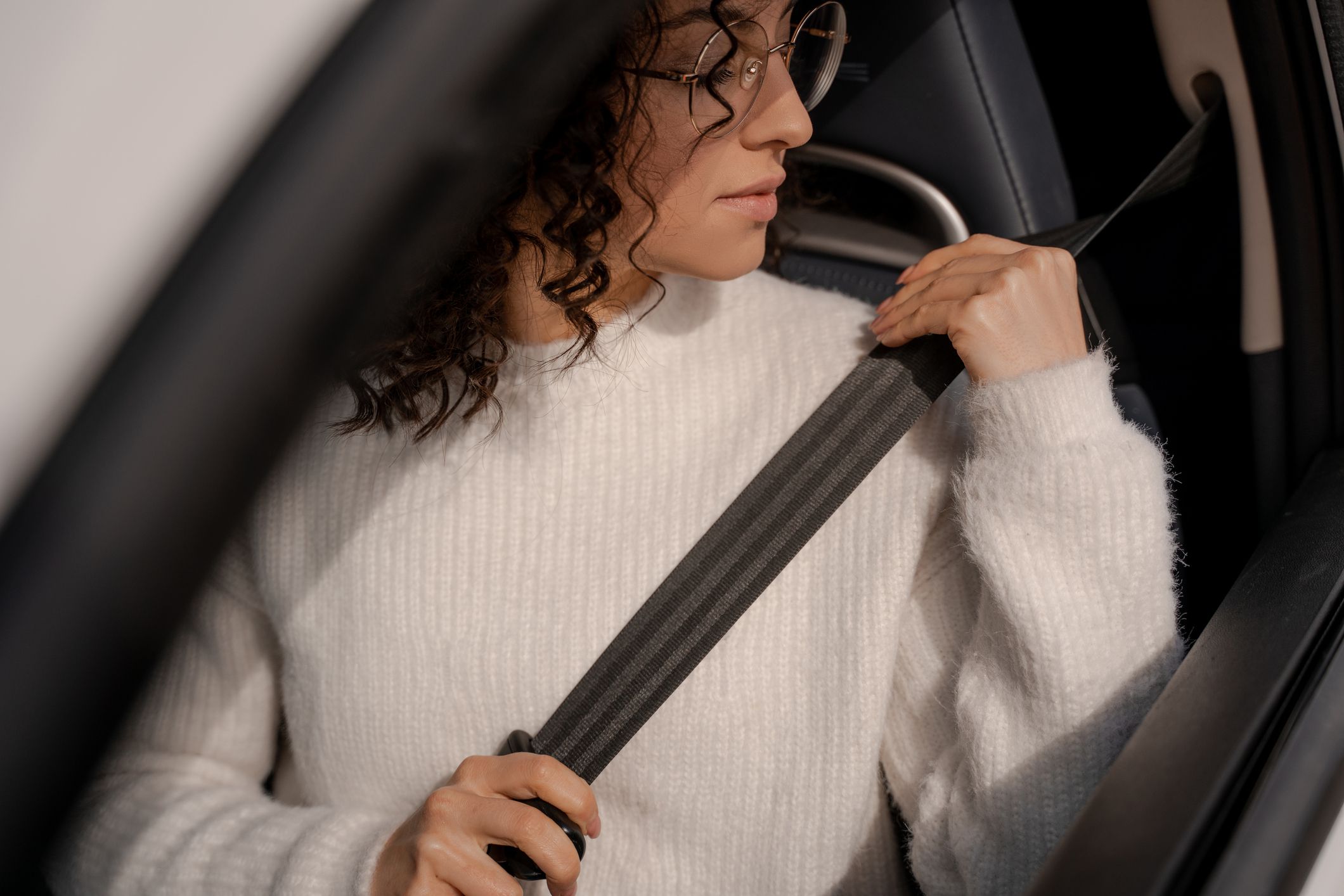 European girl buckles down seat belt in car. Concentrated young curly woman wearing glasses. Concept of driving car