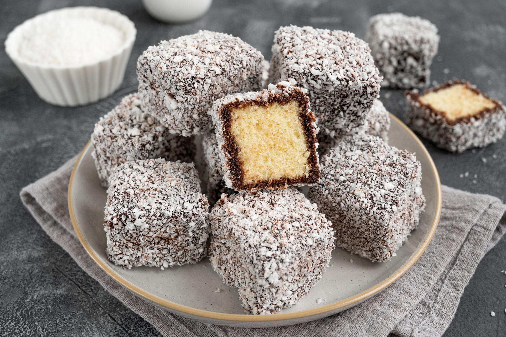 Traditional Australian Lamington cake in chocolate glaze and coconut flakes on a plate on a gray concrete background with a cup of tea. Selective focus