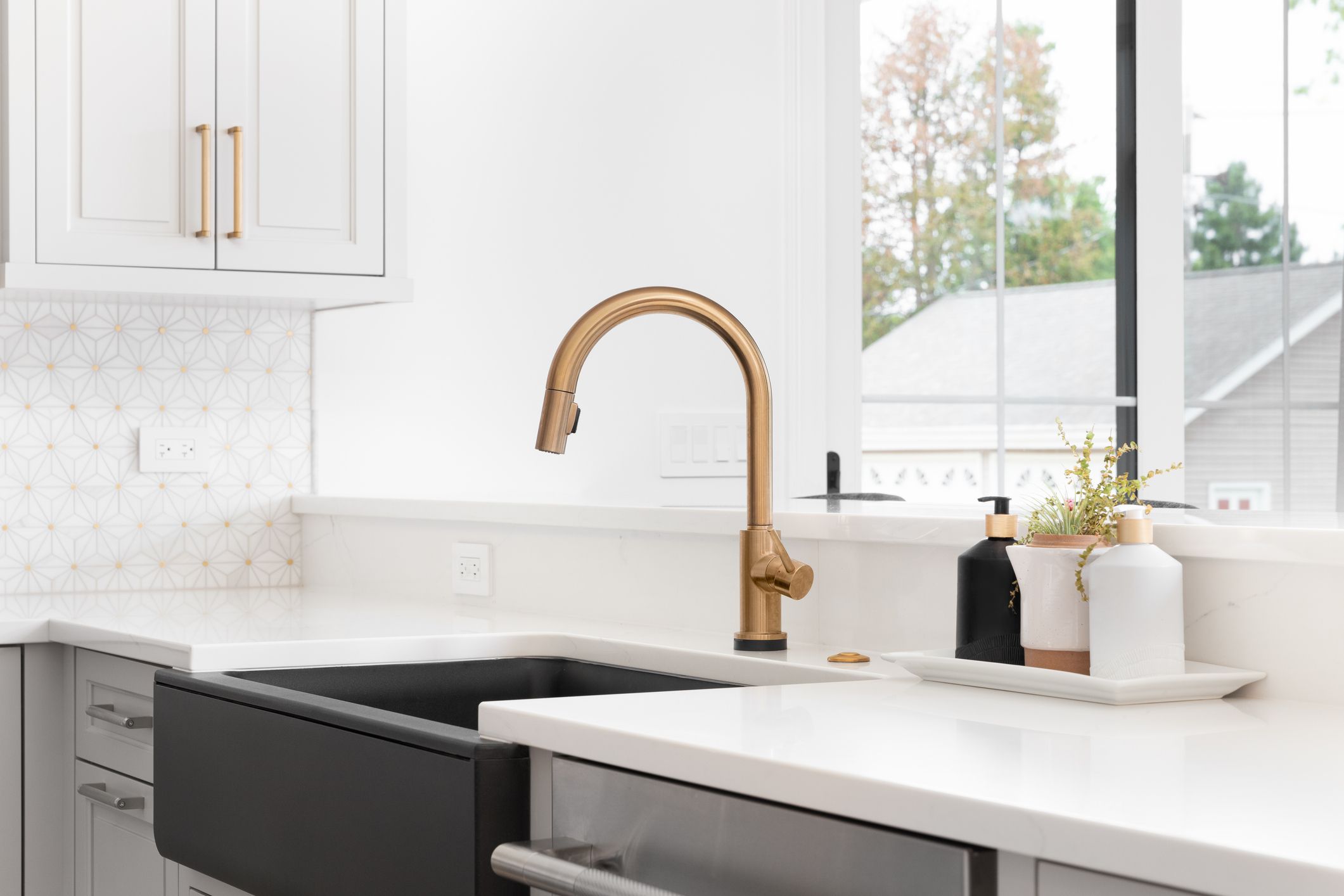 A beautiful sink in a remodeled modern farmhouse kitchen with a gold faucet, black farmhouse sink, white granite, and a tiled backsplash. No labels.