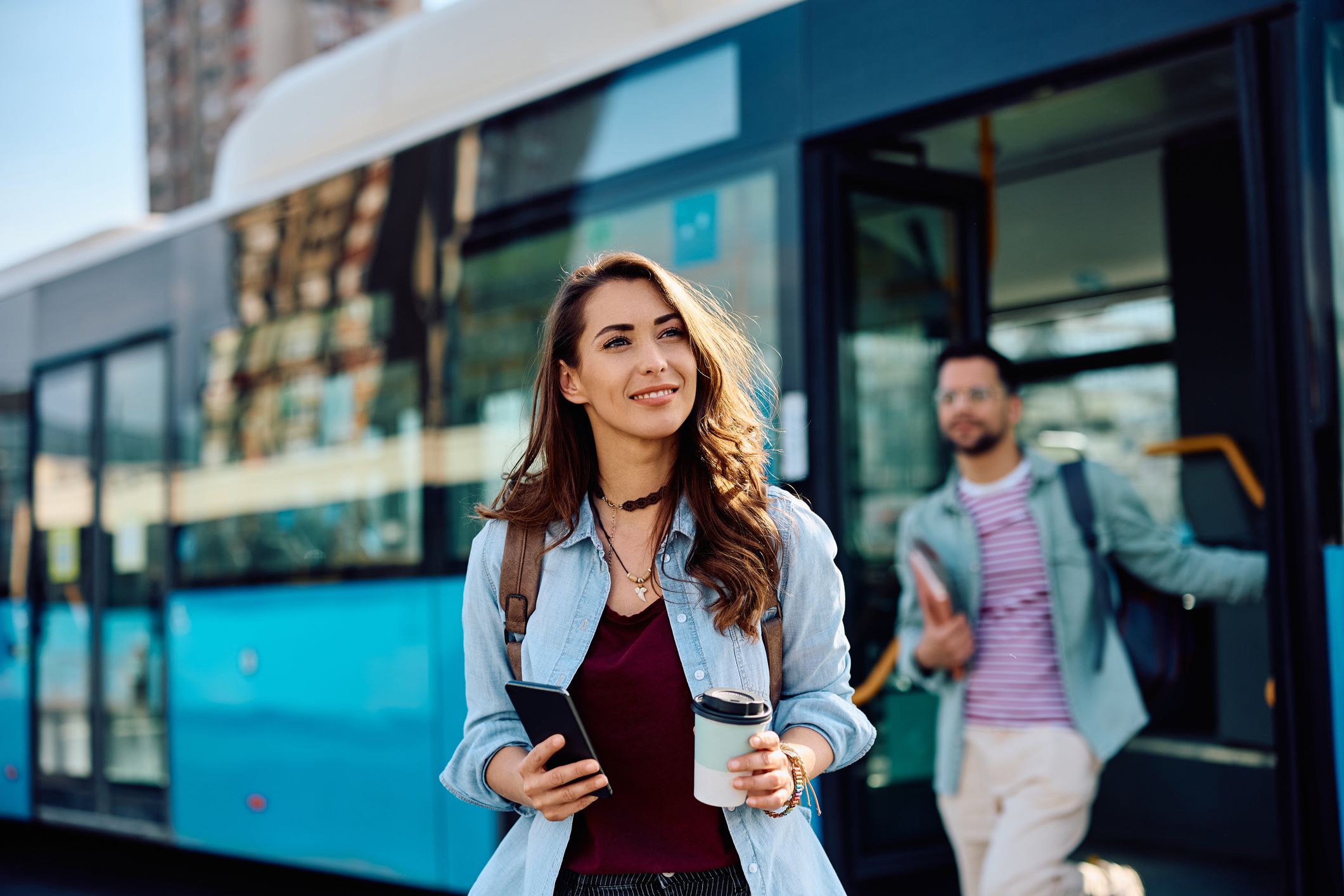 Young woman carrying coffee to go and mobile phone while getting of a bus at the station.