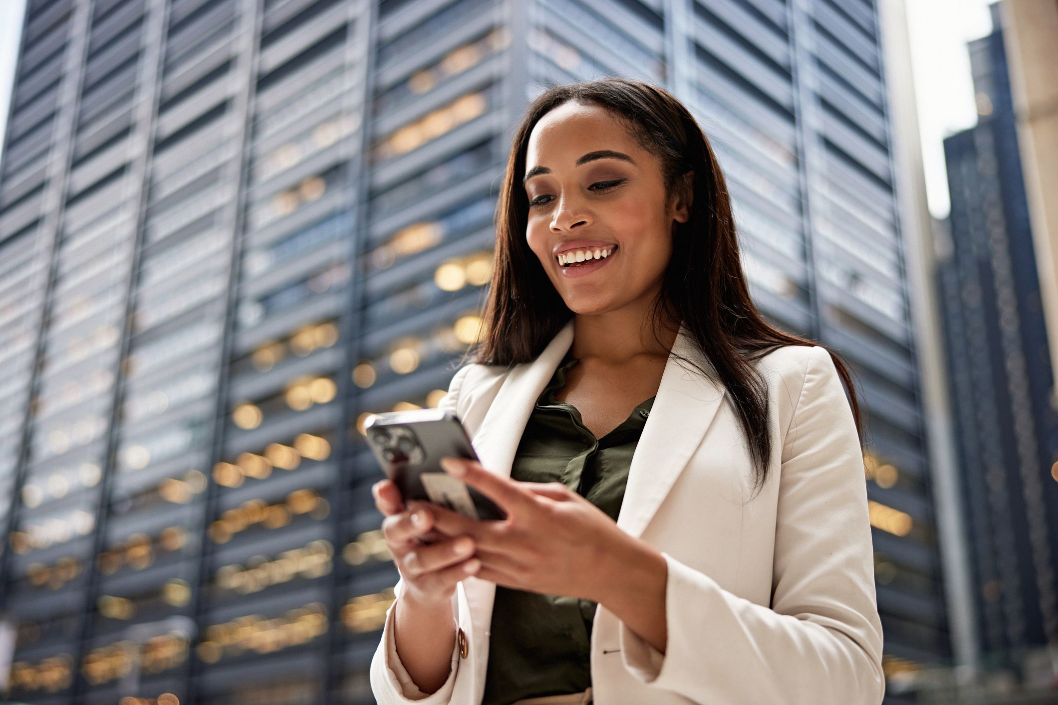 Close waist up view of longhaired Black professional in late 20s wearing office attire and communicating on portable device, NYC office building in background.