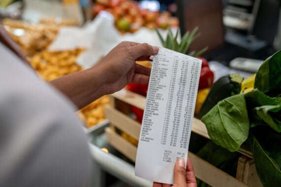 A person holds a long grocery receipt in front of a crate filled with fresh vegetables, including zucchini and spinach, carefully managing their food budget at a supermarket checkout counter.