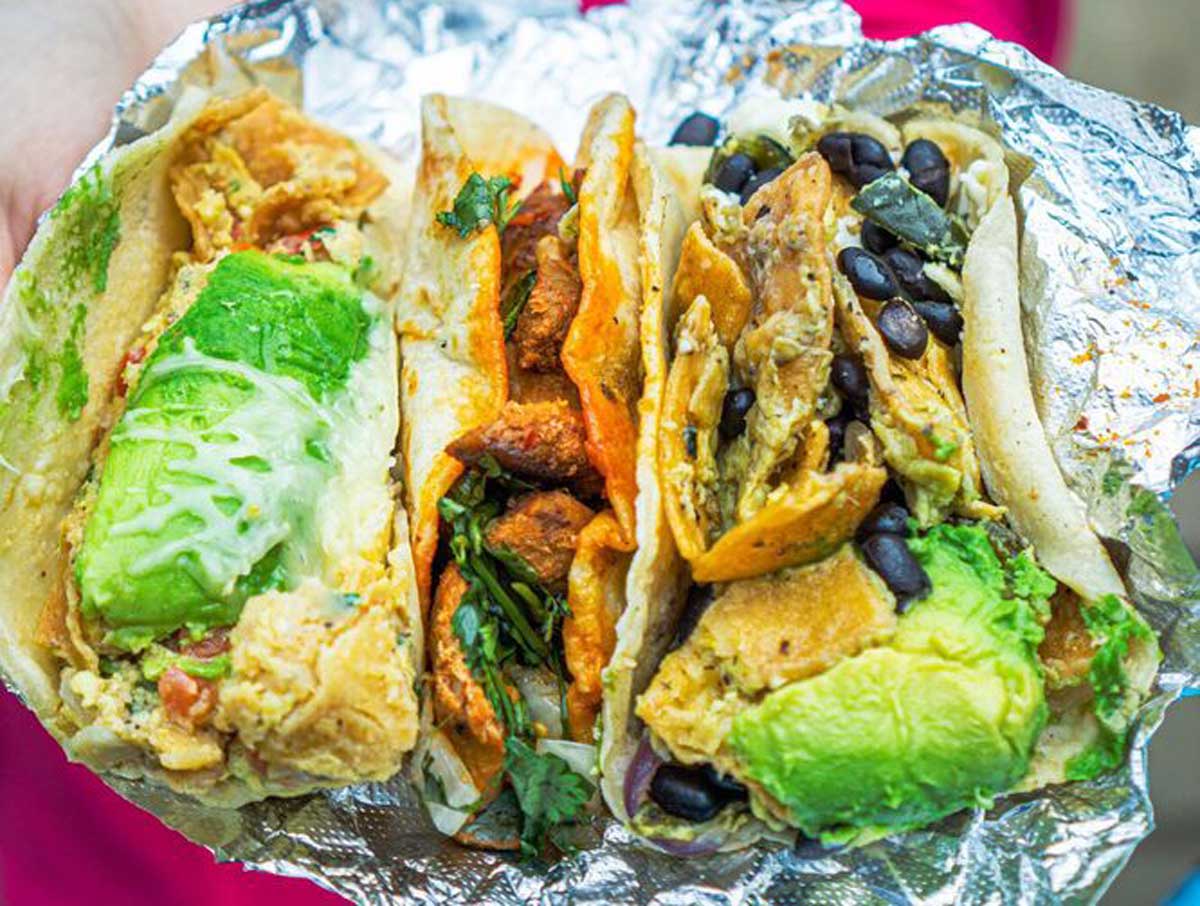Three tacos on foil, filled with avocado slices, black beans, cilantro, tortilla chips, and mixed vegetables—these could rival the best breakfast foods in every state. They're held in one hand against a blurred pink background.