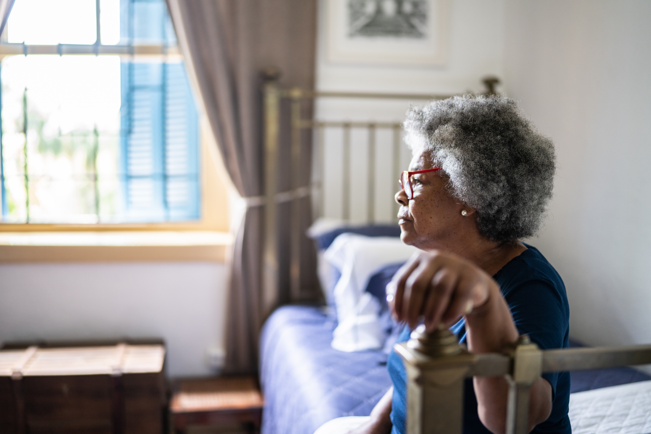 Side-View of Distraught Senior Woman Sitting on Her Small Bed, Selective Focus