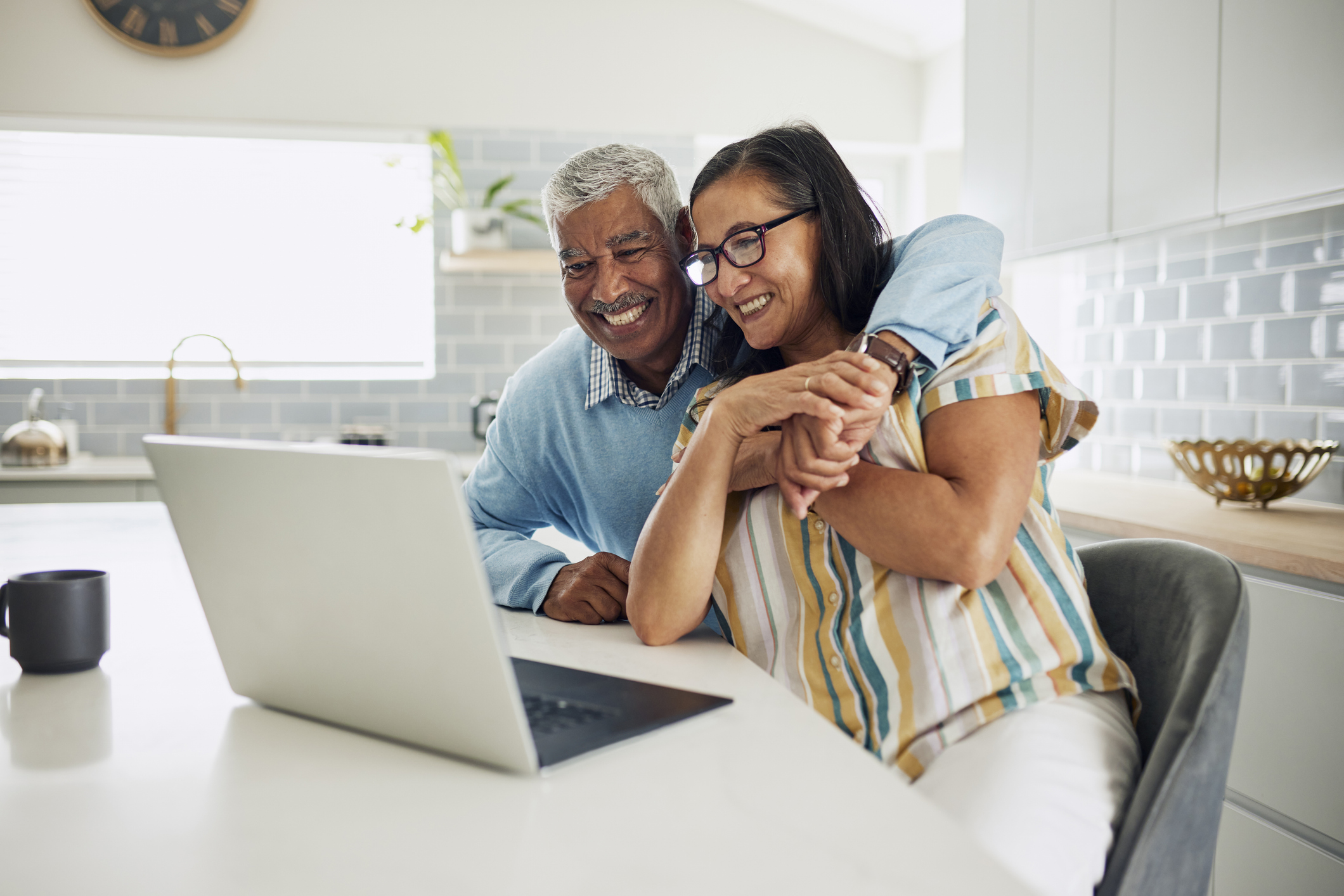 Senior Couple Hugging in the Kitchen While Looking at a Laptop