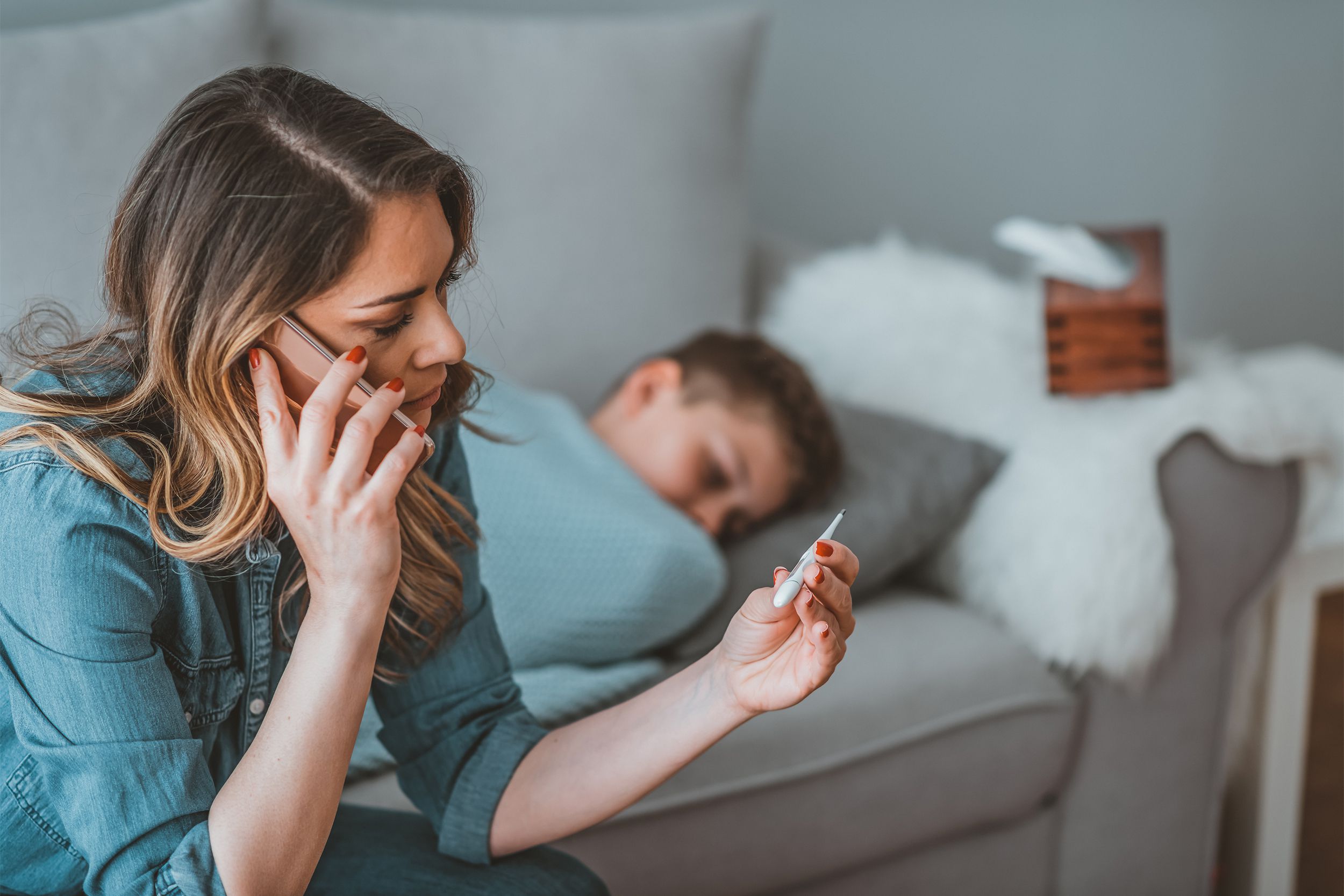 Mother on the phone looking at thermometer with sick kid in the background