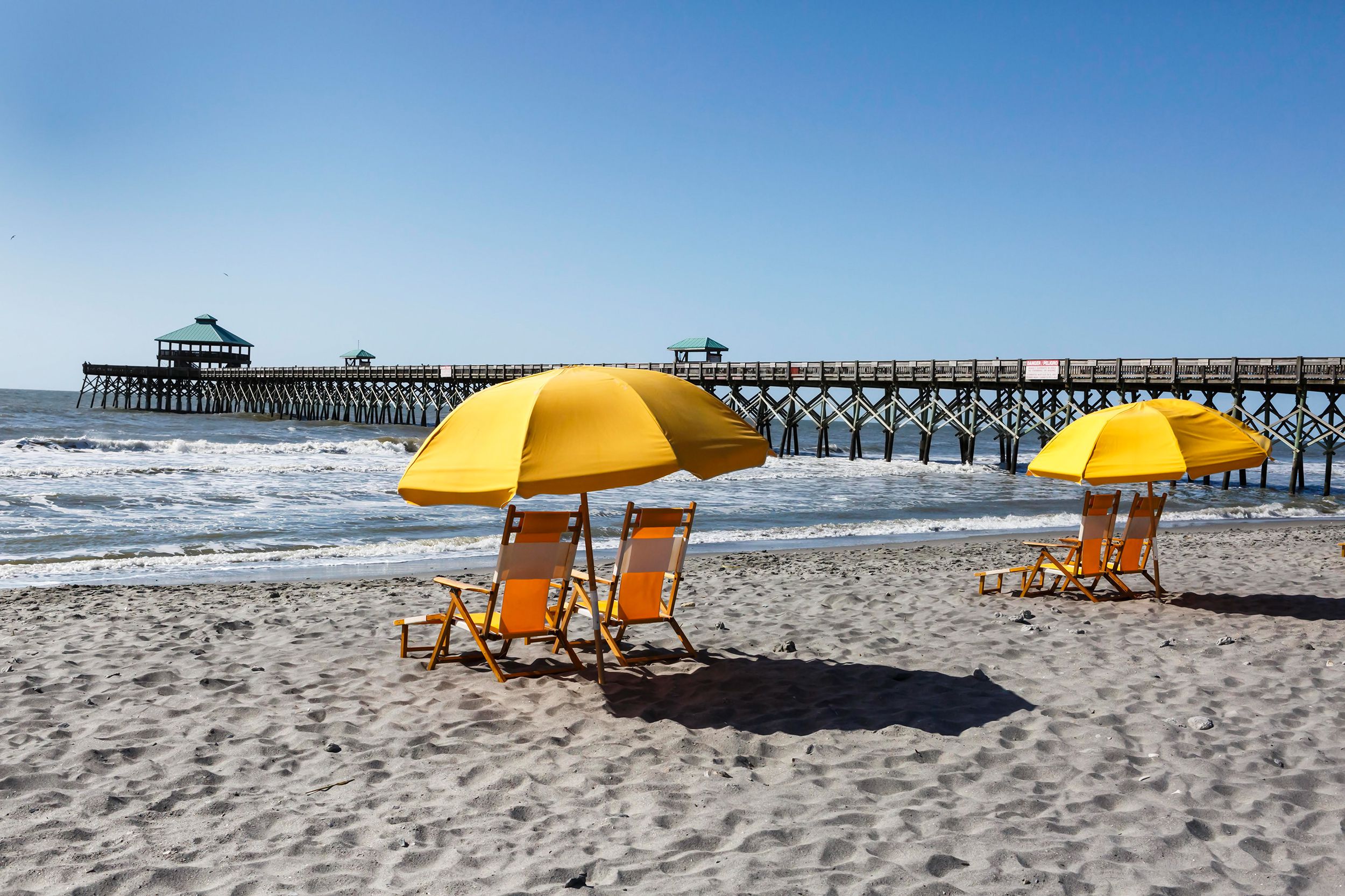 Two sets of yellow empty beach chairs with an umbrella in the sand and the pier at Folly Beach, South Carolina during the day