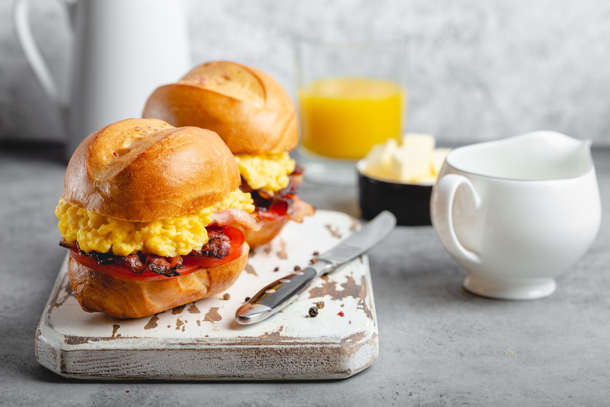 Breakfast sandwiches with scrambled egg, bacon, cheese, tomato on white wooden board, glass with fresh orange juice, white background. Making breakfast concept, selective focus