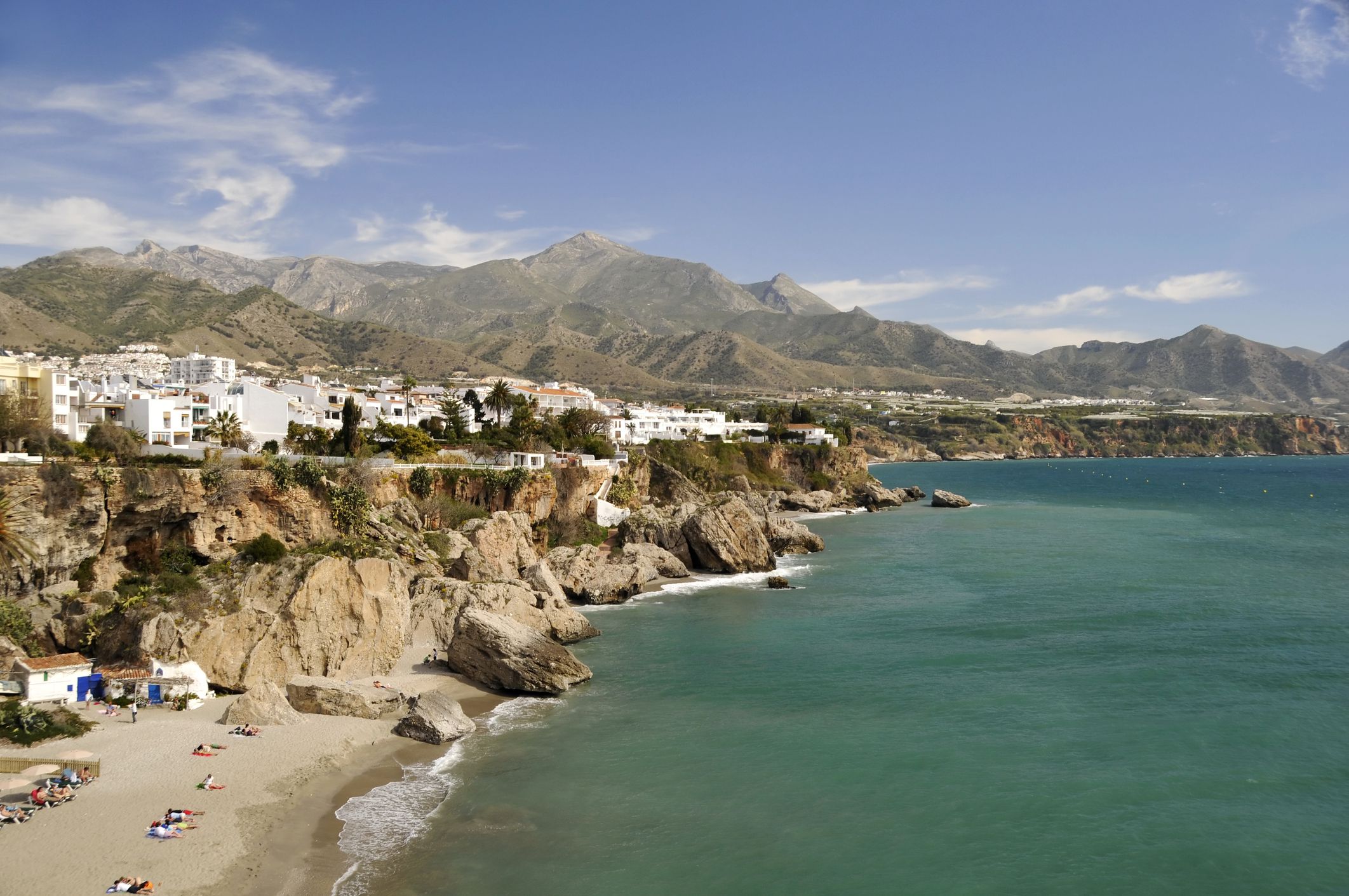 The view of the Nerja in Malaga, Spain, is pictured. There are mountains and an outcropping of rock to the left of the sea dotted with white and pale-colored houses. The sea is teal and blue with a blue sky and wispy white clouds in the background.