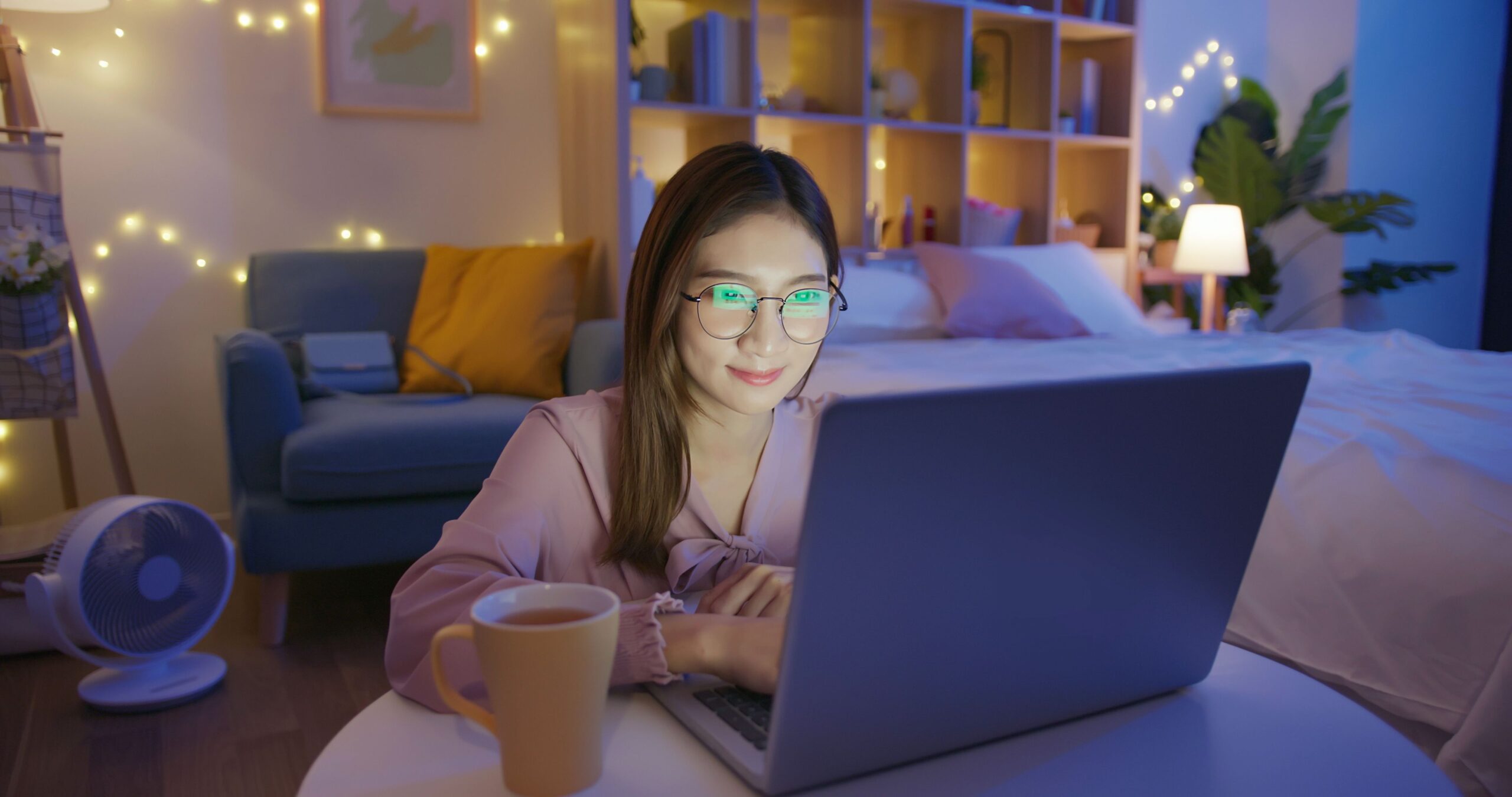 asian freelancer woman wearing eyeglasses works by laptop with tea at home in the evening