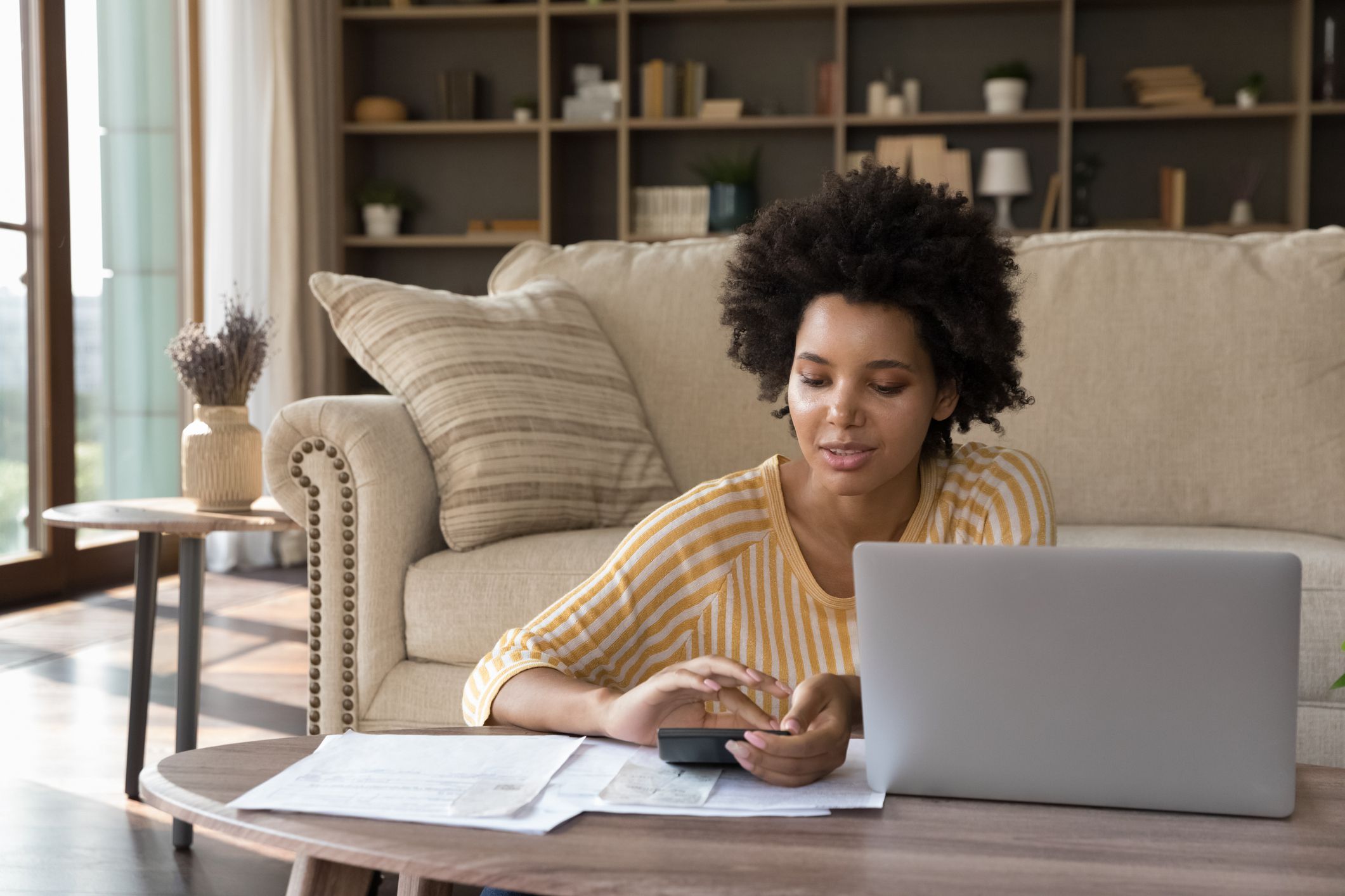 African woman makes calculations on calculator pay utility bills, loan via e-bank application on computer. sit in living room alone. Personal family budget management, modern tech, accounting concept