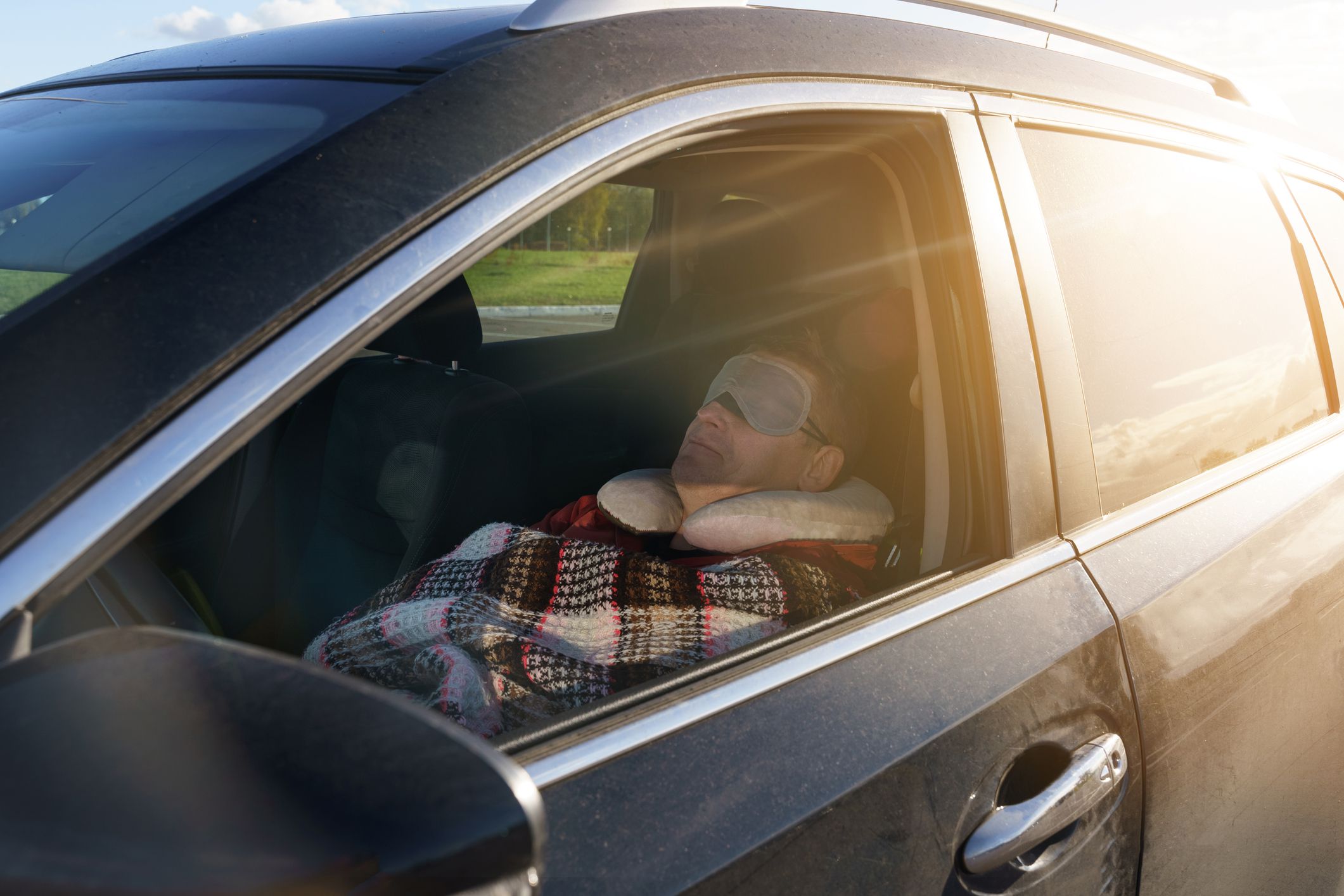 A tired driver sleeps in the car during the day, covered with a blanket and wearing a sleep mask over his eyes. Security concept.