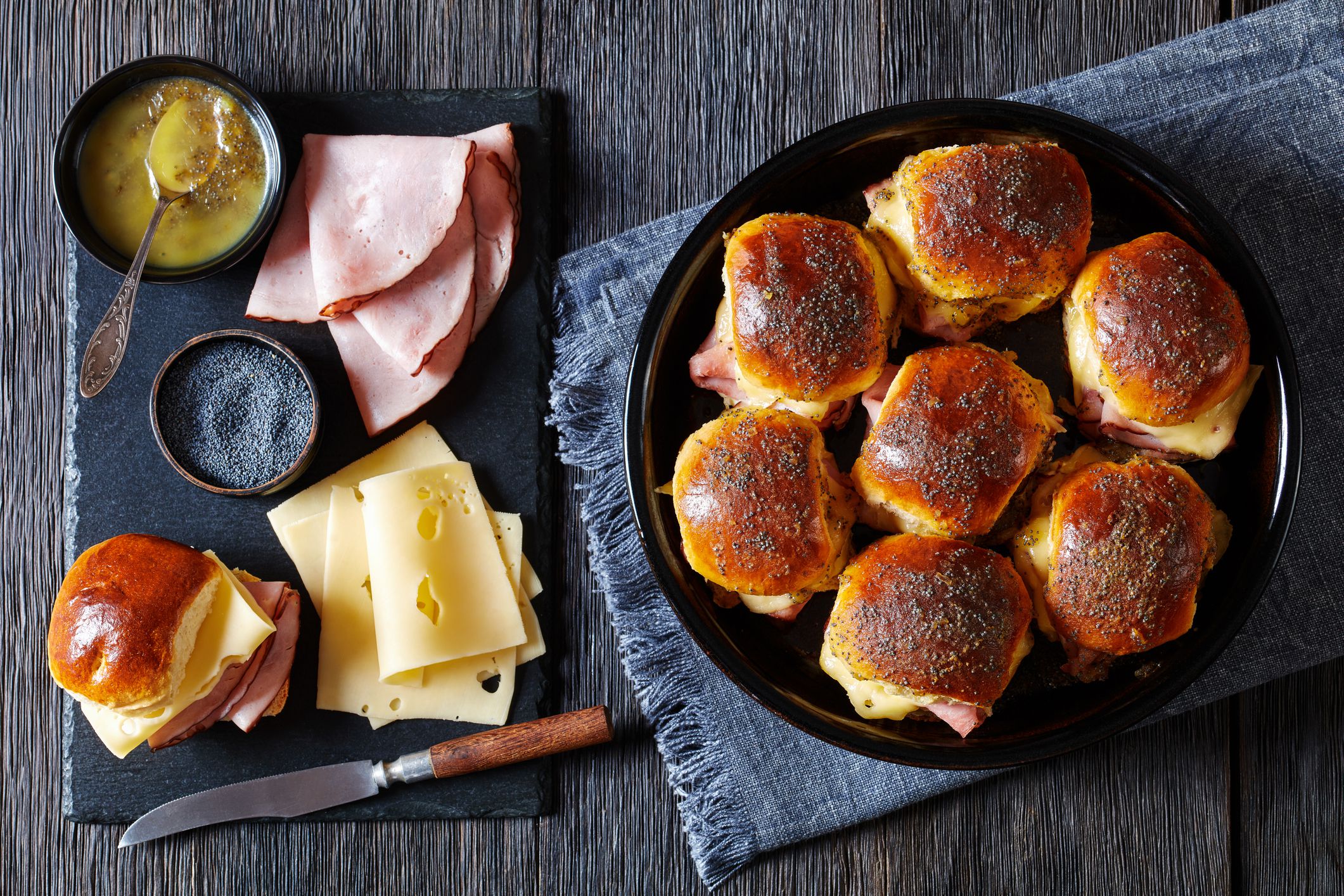 Hawaiian Ham and Swiss Sliders in baking dish with ingredients on black stone board on dark wood table, horizontal view from above, flat lay