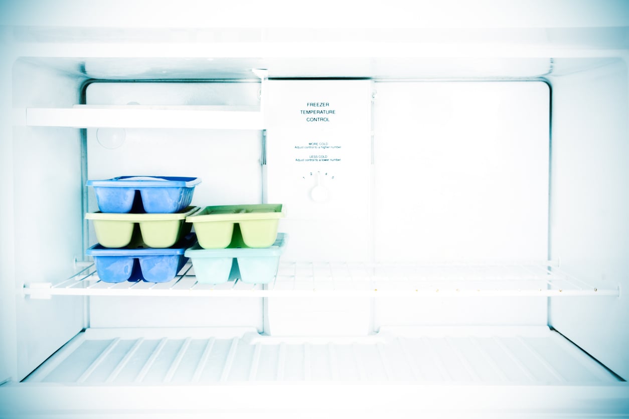 An open, mostly empty freezer with three ice cube trays—two blue and one green—stacked on the left side of the middle shelf. The freezer temperature control is visible at the back.