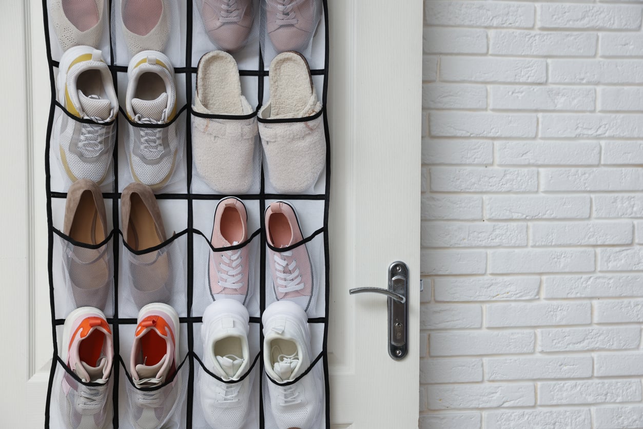 A white door with a hanging shoe organizer holds six pairs of shoes and slippers, with a silver door handle visible. The background shows a white brick wall.