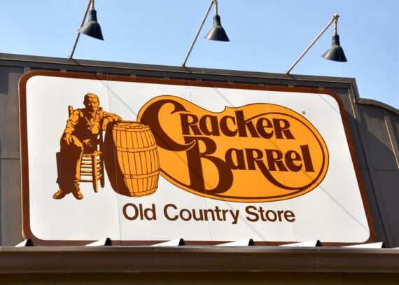 A large Cracker Barrel Old Country Store sign shows a man sitting in a chair beside a barrel, with the restaurant’s name in bold orange and brown letters on a white background.