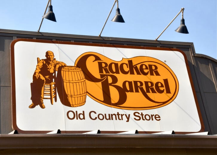 A large Cracker Barrel Old Country Store sign shows a man sitting in a chair beside a barrel, with the restaurant’s name in bold orange and brown letters on a white background.
