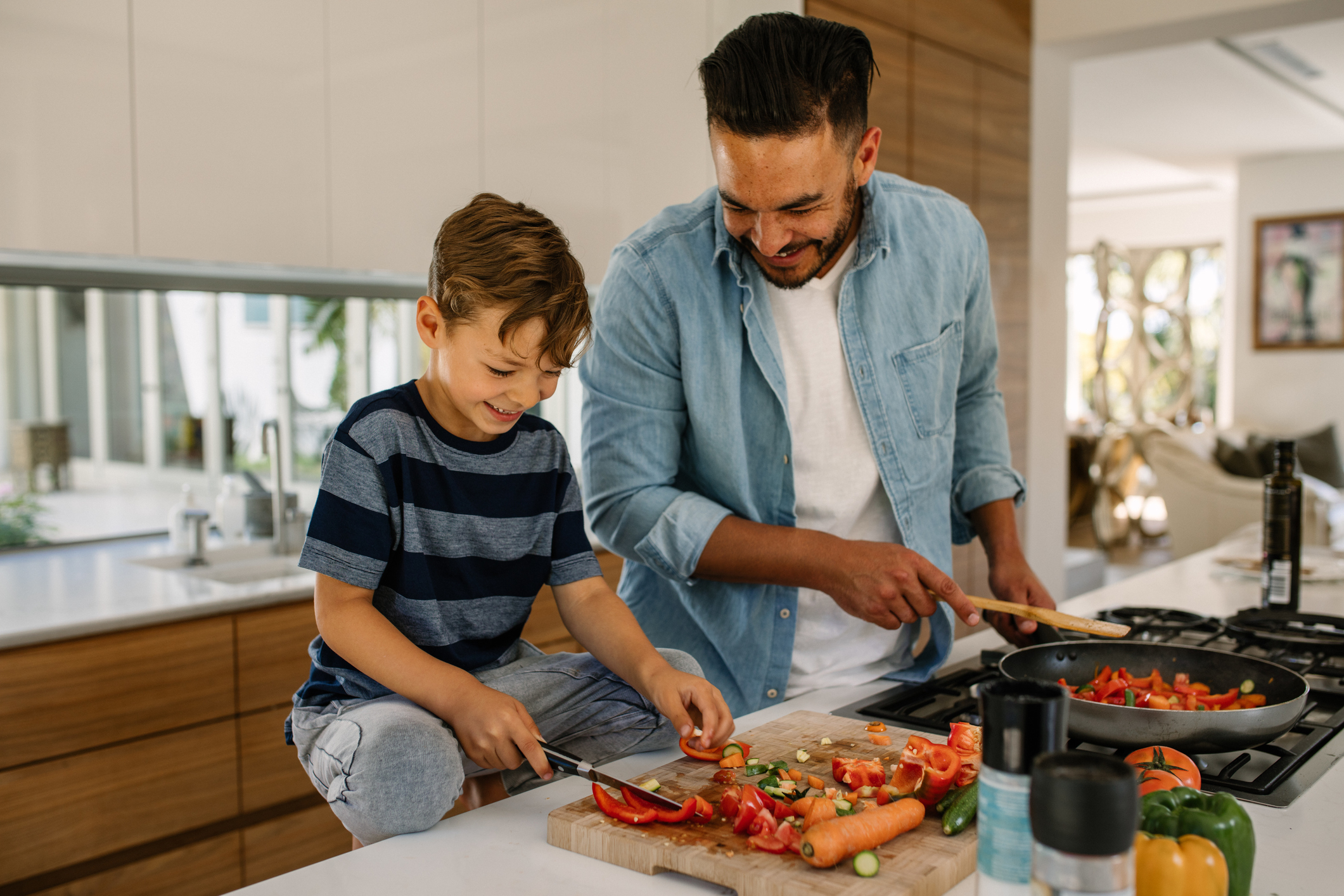 Laughing Father and Son Cooking, Son Is Cutting Up Vegetables, Father Is Using a Skillet