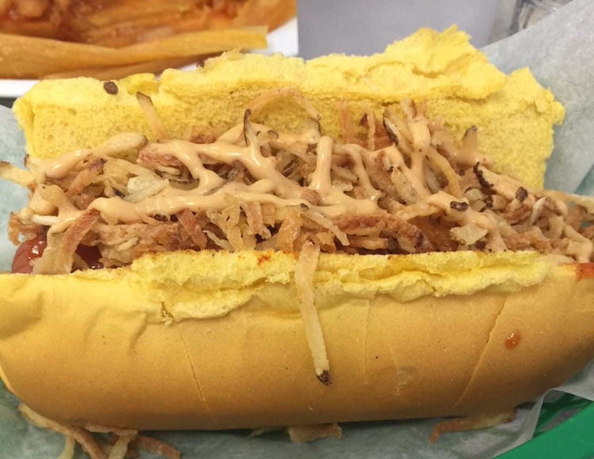 A hot dog in a yellow bun topped with crispy fried onions and a drizzle of creamy sauce, served on wax paper—just like those found at under-the-radar hot dog stands. In the background, there are fries and a drink.