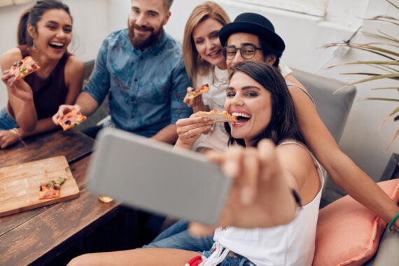 A group of five friends sit closely together on a couch, smiling and eating pizza while one person in front takes a selfie with a smartphone. They appear happy and relaxed, enjoying each other's company.