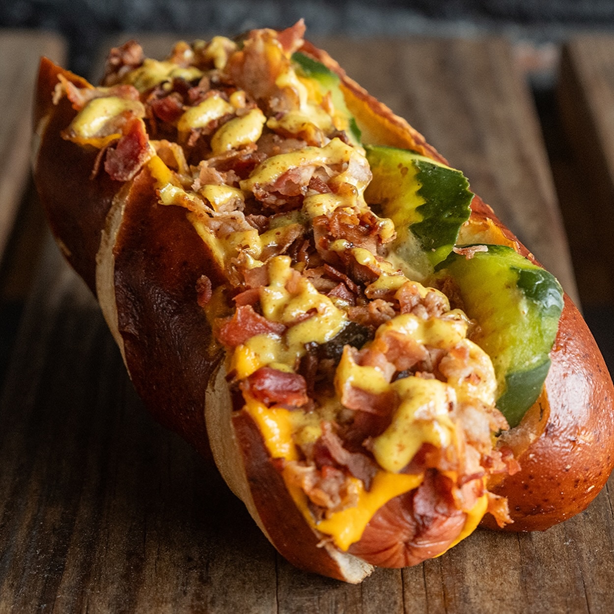 A gourmet hot dog in a pretzel bun topped with melted cheese, chopped bacon, sliced pickles, and a drizzle of mustard—much like the creations from under-the-radar hot dog stands—served on a rustic wooden surface.
