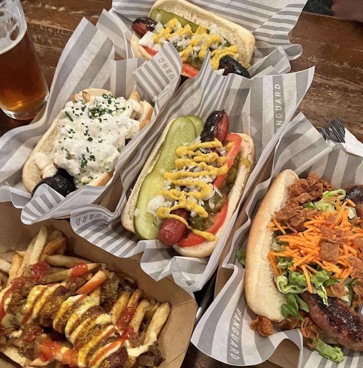 Five gourmet hot dogs with creative toppings and a serving of loaded fries sit next to a glass of beer, showcasing flavors found at the best under-the-radar hot dog stands—each dog topped with pickles, onions, mustard, vegetables, and cheese.