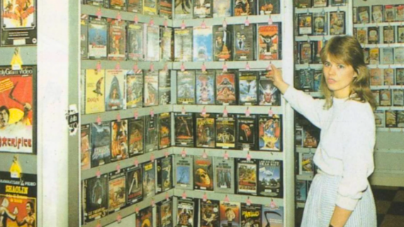 A woman in a light outfit stands in a video rental store, pointing at shelves filled with VHS tapes in colorful covers. Surrounded by racks packed with various movie titles, she seems to be searching for something special among these analog treasures&mdash;perhaps reminiscing about the days before digital things took over home entertainment.