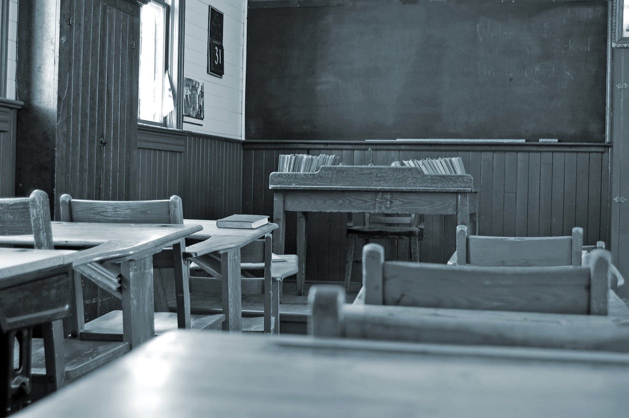 Black-and-white photo of an empty classroom with wooden desks and chairs, books stacked on the teacher’s desk—a nostalgic setting where facts boomers learned in school or even debunked myths might have once filled the chalkboard at the front.