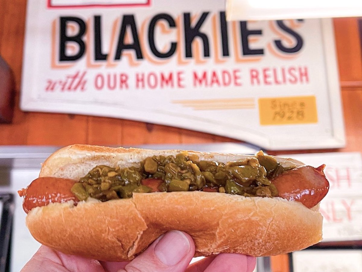 A hand holds a hot dog topped with green relish in front of a vintage sign reading "BLACKIE'S with OUR HOME MADE RELISH," capturing the charm of under-the-radar hot dog stands inside a nostalgic restaurant.