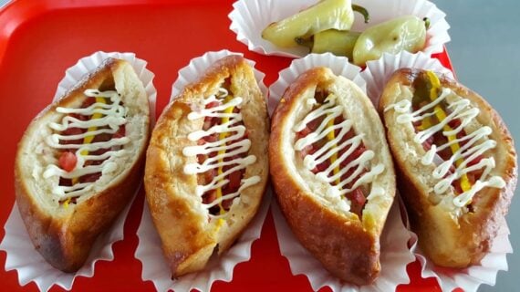 Four oval-shaped baked pastries inspired by under-the-radar hot dog stands, filled with diced vegetables and topped with zigzagged mayonnaise, served on a red tray with a side of pickled green peppers.