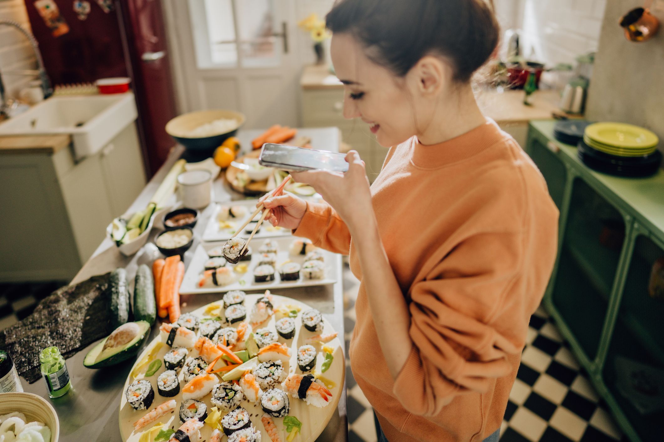 Photo of a smiling female food blogger photographing sushi plate and holding a pair of chopsticks by the kitchen counter.