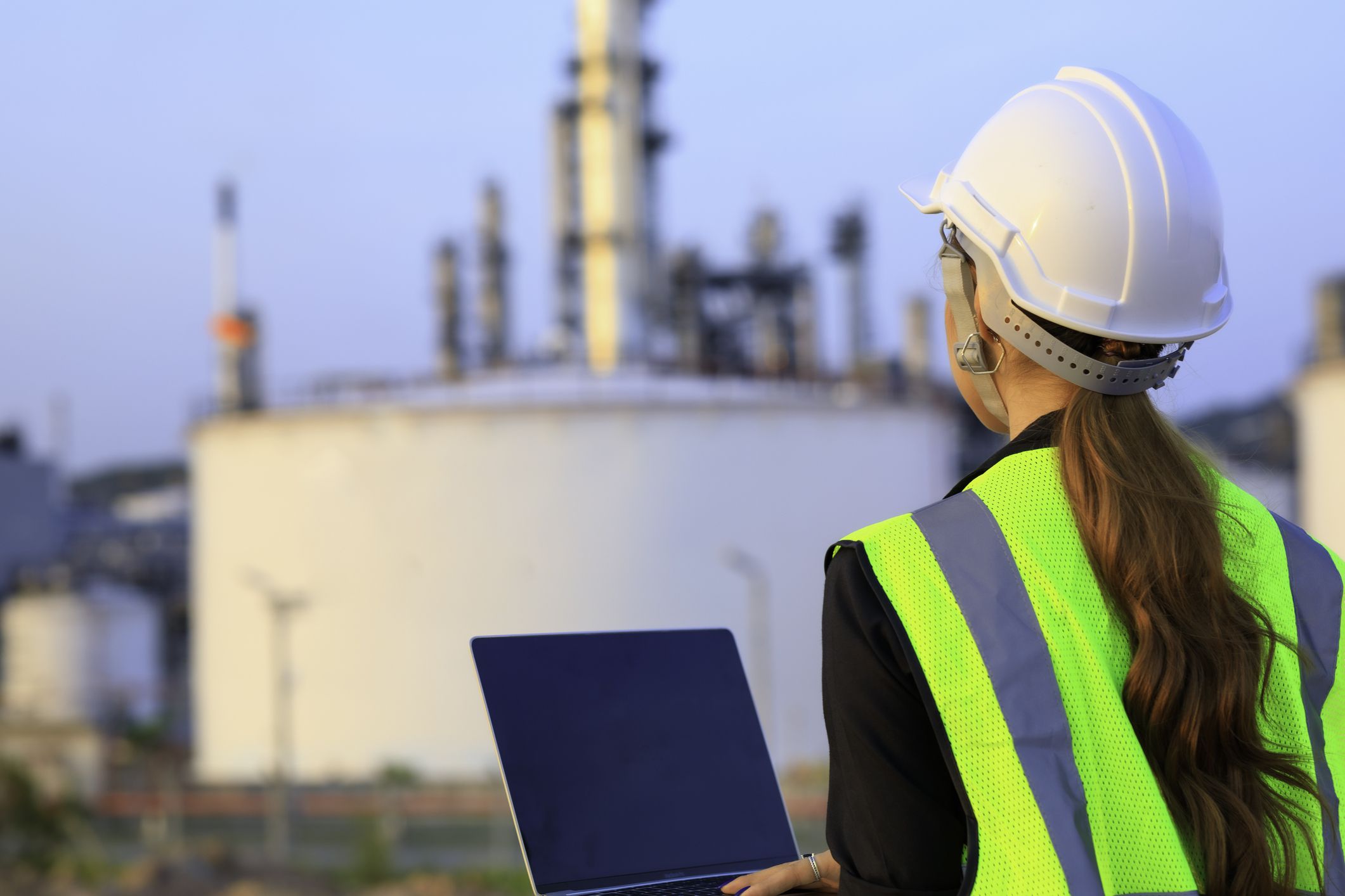 Female engineer foreman wearing safety helmet using laptop working in oil refinery industry plant background.