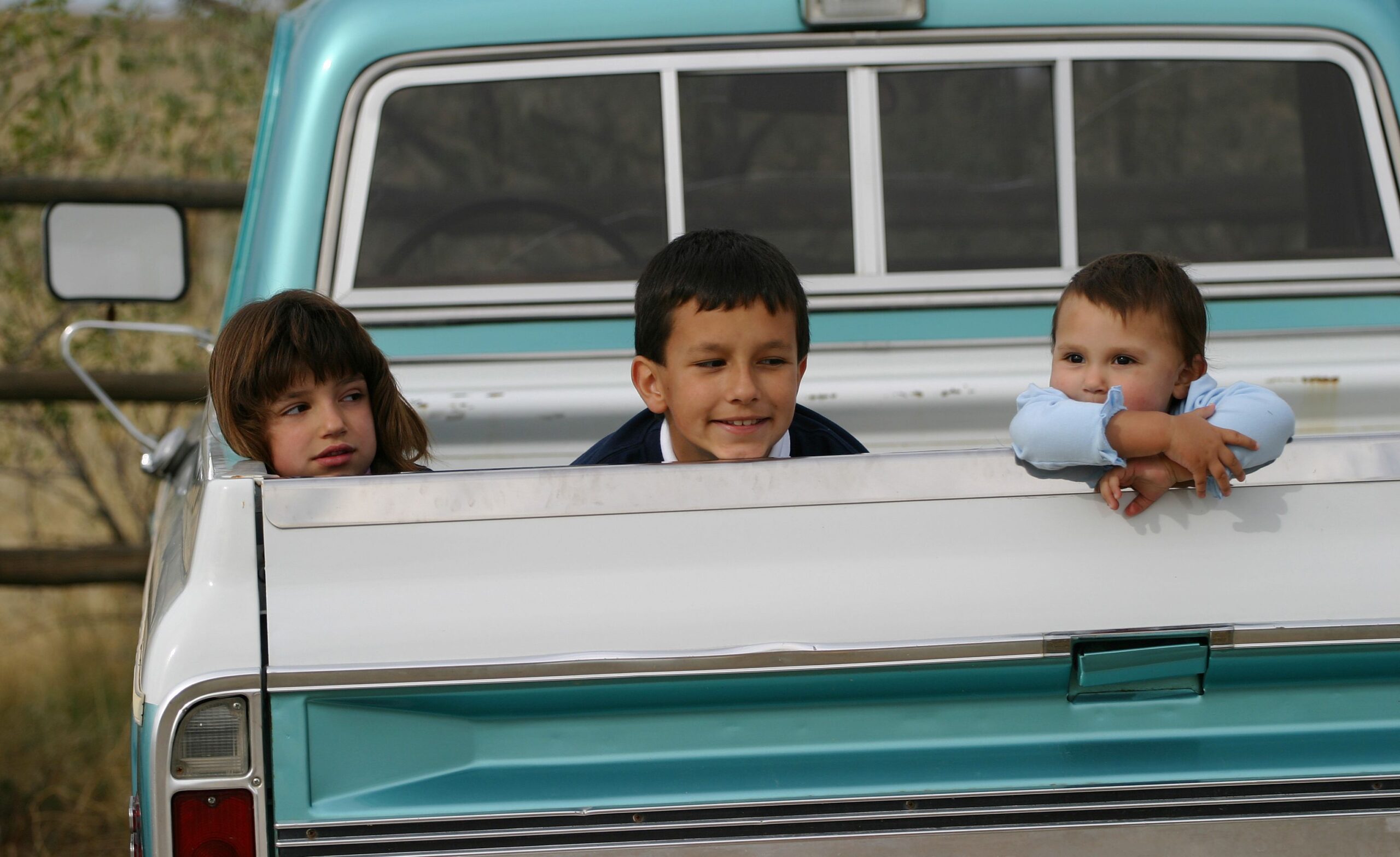 Three Hispanic American children, each with different expressions, hanging out in the back of a pick up truck. Siblings.