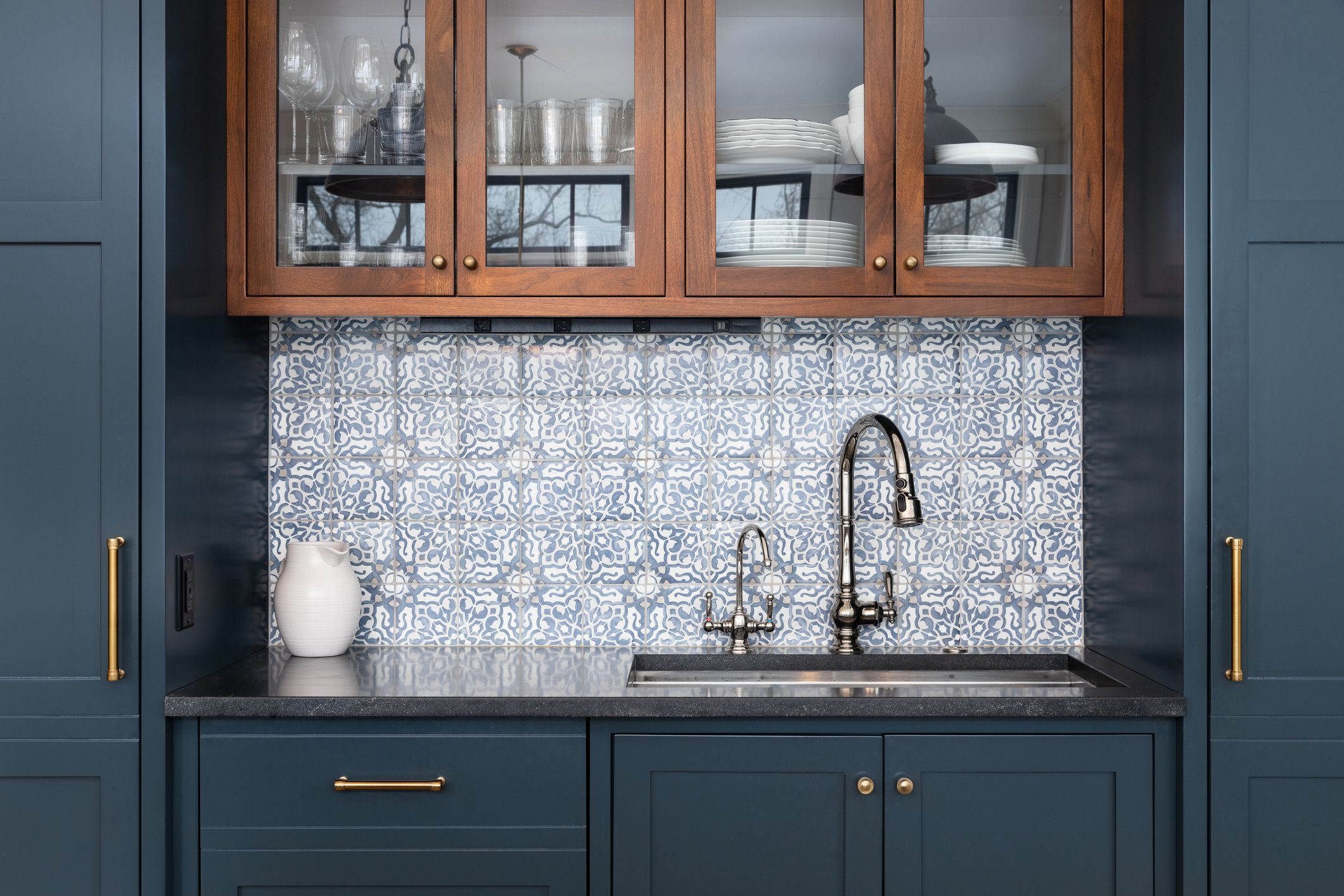 A kitchen sink with a beautiful pattern tiled backsplash with a chrome faucet, black granite countertops, and surrounded by blue and wood cabinets.