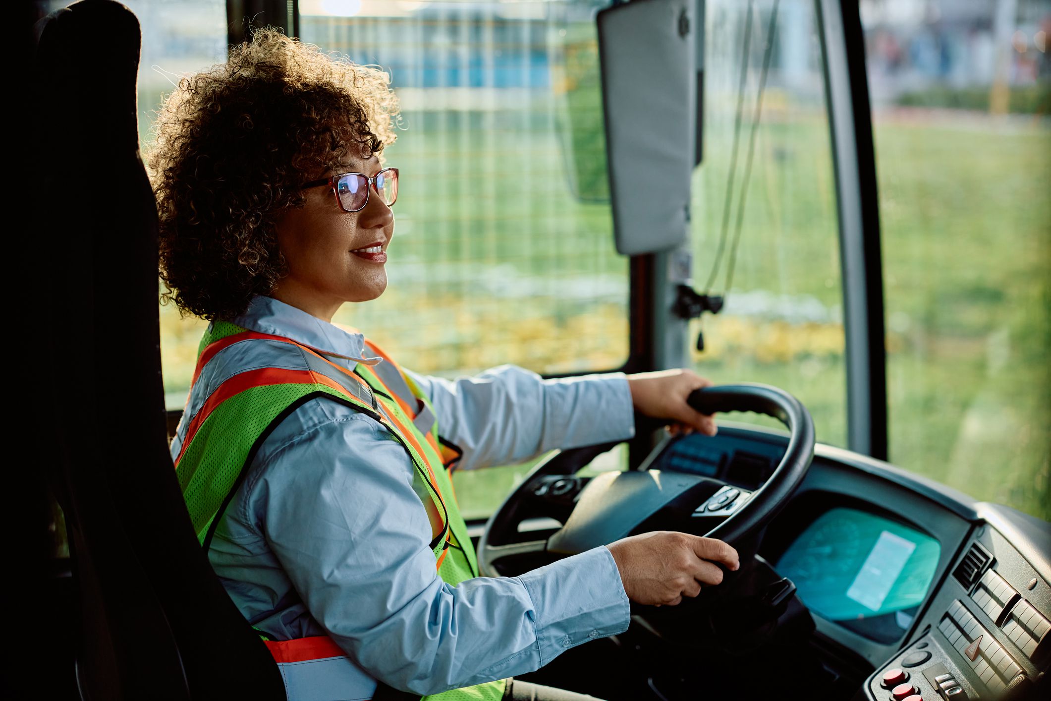 Happy female bus driver looking at rear view mirror while parking in reverse at the station.
