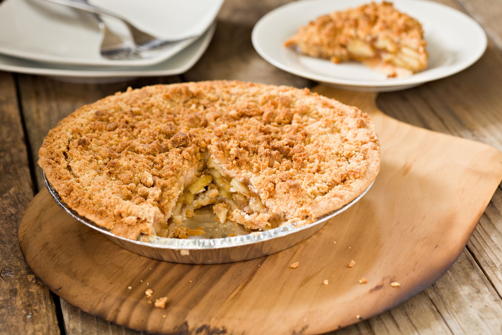 A high angle close up shot of a freshly baked dutch apple pie sitting on a wooden pizza peel and a serving of pie in the background. Shot on a grungy old wooden table.