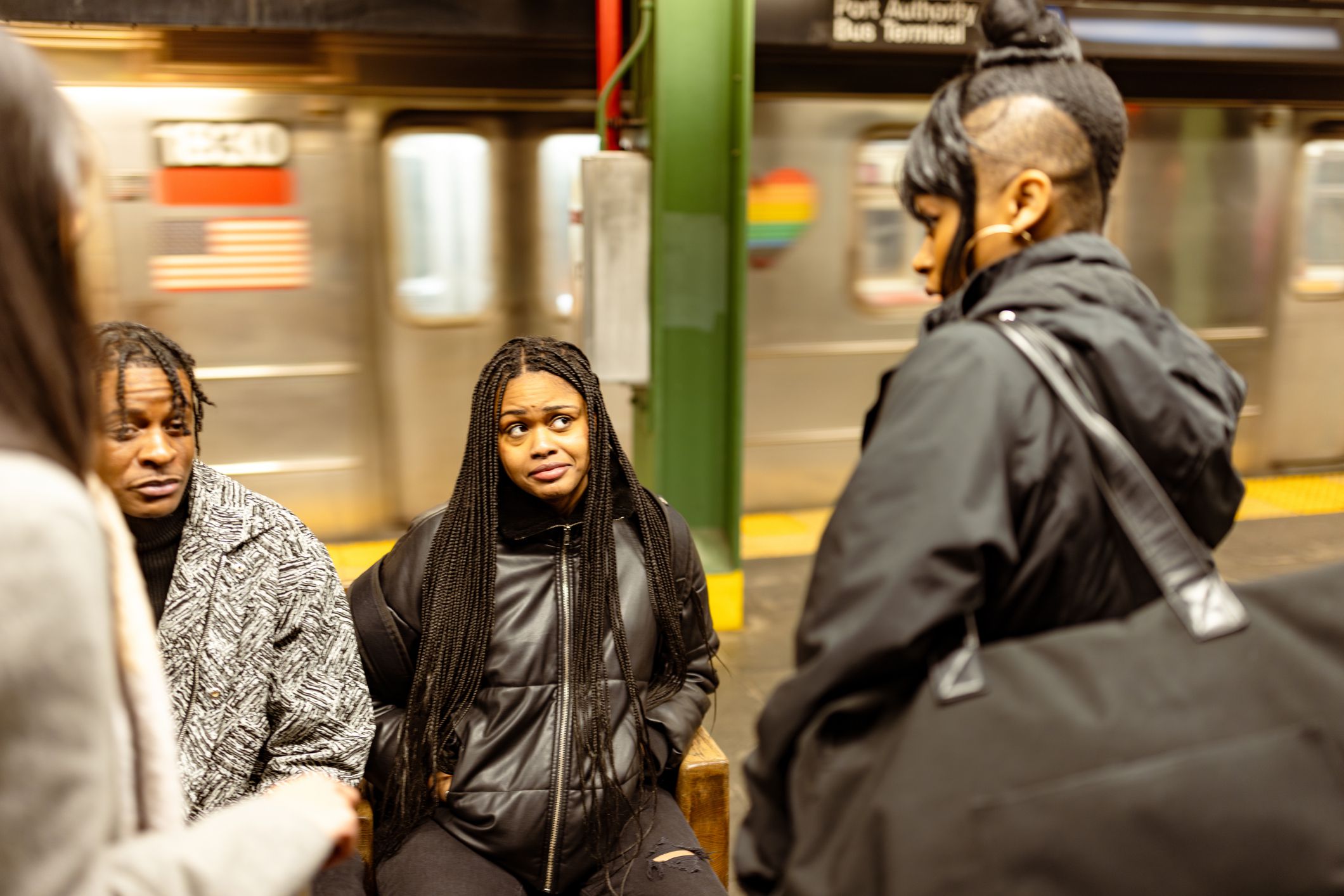Group of diverse young students engages in conversation while sitting and standing at a bustling New York subway station, dressed in casual attire, with backpacks hinting at a day of learning.