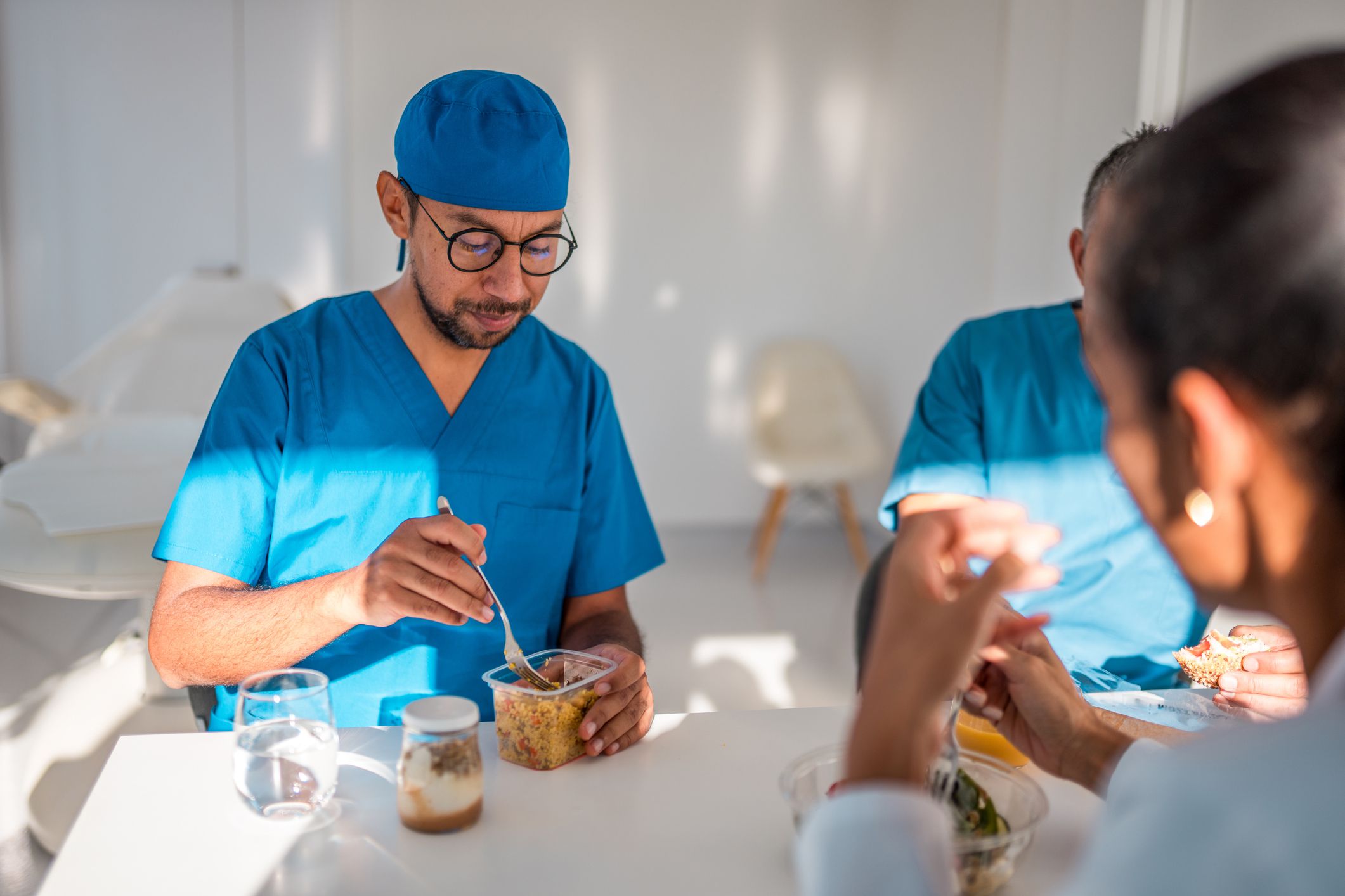 A male doctor in blue scrubs eats lunch while conversing with female colleagues in a brightly lit hospital break room, depicting a moment of rest in a medical environment.