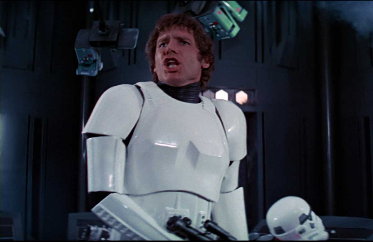 A man in white stormtrooper armor stands in a futuristic control room, looking startled and talking. A stormtrooper helmet lies on the console in front of him. The background includes dark walls and control panels.