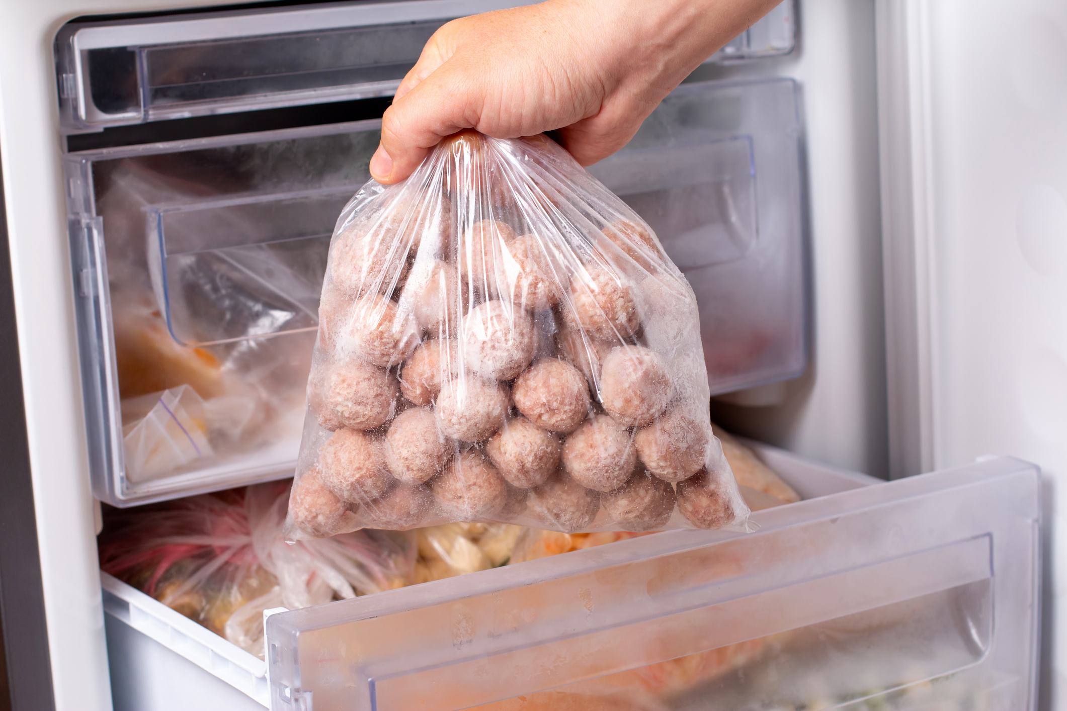 Hand Holding a Bag of Frozen Meatballs Over Bottom Opened Freezer Door