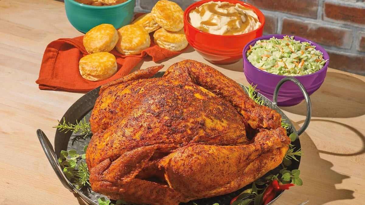 A whole Popeyes Thanksgiving turkey on a platter with herbs, surrounded by bowls of coleslaw, mashed potatoes with gravy, biscuits, and another side dish on a wooden table.