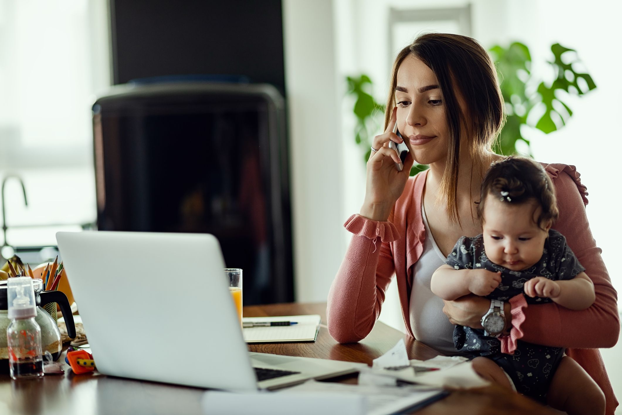 Working mother with baby daughter communicating on mobile phone while reading an e-mail on the computer.